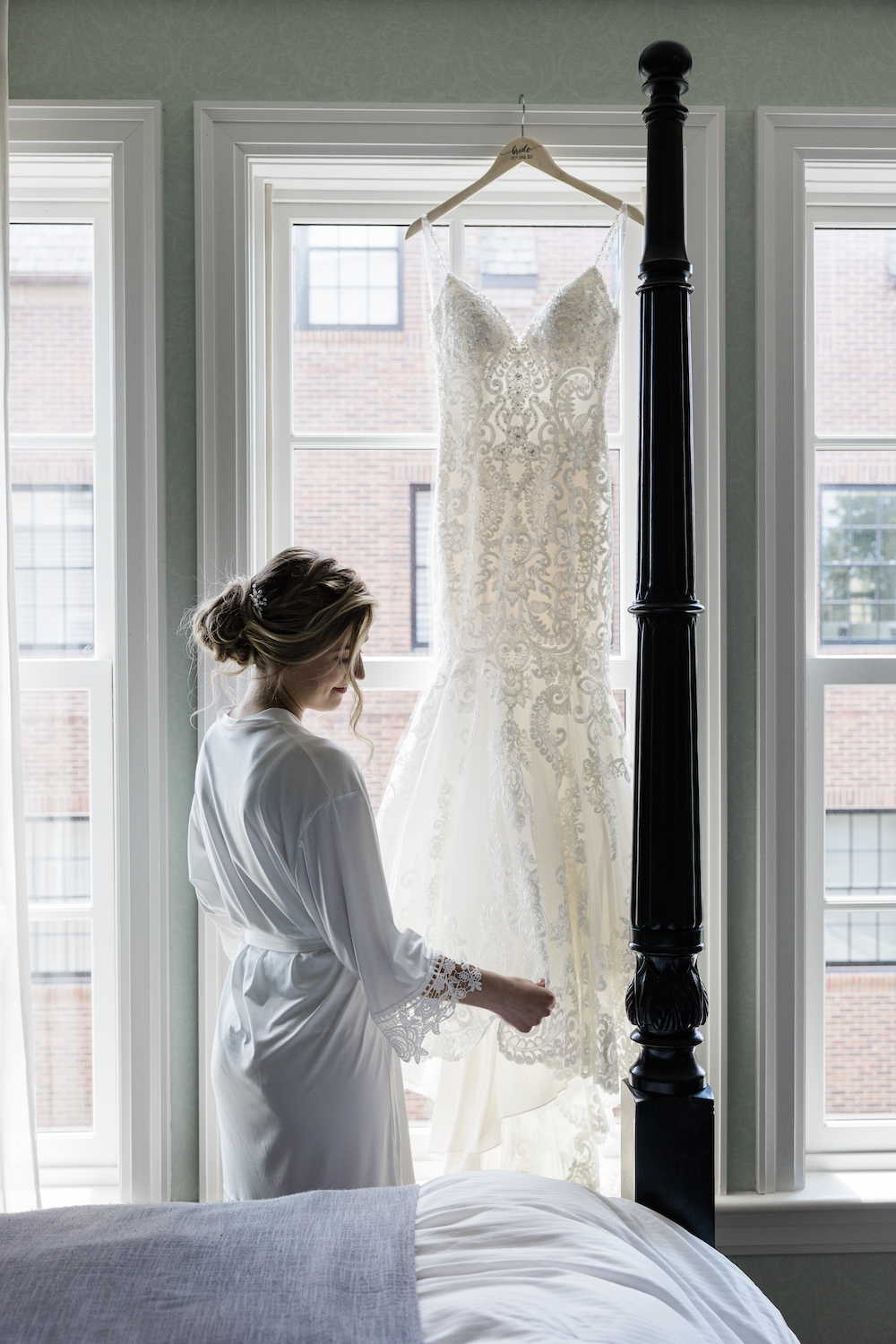 Bride in her robe touching the fabric of her wedding dress hung from the window of her suite, captured by Holly Green Photography